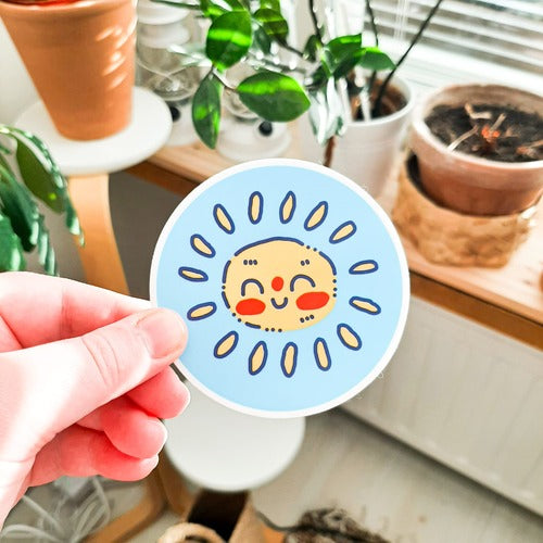 Kuvassa on henkilön käsi pitelemässä tarraa, joka on iloinen aurinko. Tarra on pyöreä ja sillä on sininen tausta. Kuvan taustalla on kodin huonekaluja ja kasveja.