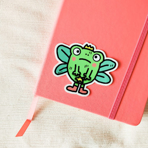 Kuvassa on sammakkotarra. Sammakolla on surullinen ilme ja silmät hieman kierossa. Sillä on myös keijun siivet, taikasauva, kruunu ja vaaleanpunaiset sukat.