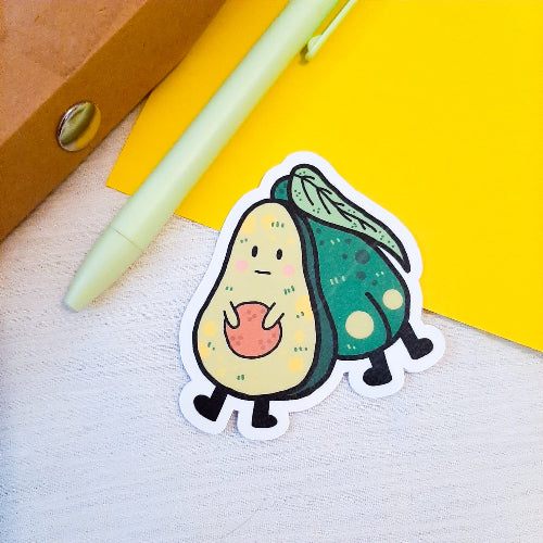 Kuvassa on vihreä tarra, jossa on kaksi avokadoa. Toisella on myrtsi ilme ja toisella näkyy pylly. Taustalla on vihreä kynä, keltaista paperia ja valkoista kangasta.