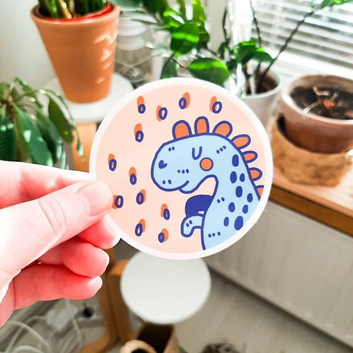 Kuvassa on henkilön käsi pitelemässä tarraa, joka on sininen dinosaurus. Tarra on pyöreä. Taustalla on kodin huonekaluja ja kasveja.