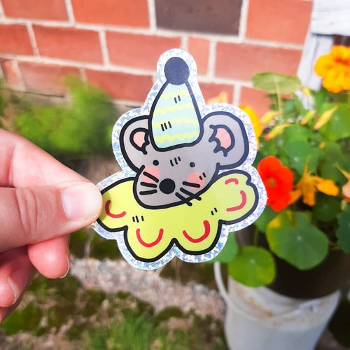 Kuvassa on henkilön käsi pitelemässä tarraa, joka on harmaa hiiri. Hiiri on pukeutunut juhlahattuun ja juhlalliseen kaulukseen. Tarra on glitterinen. Taustalla on tiiliseinä ja kasveja.