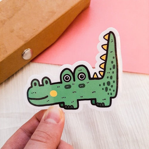 Kuvassa on henkilön käsi pitelemässä tarraa, jossa on iloinen vihreä krokotiili. Taustalla on ruskeaa, pinkkiä sekä valkoista.