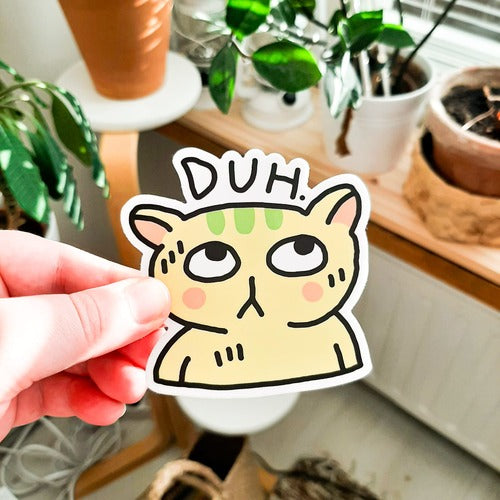 Kuvassa on henkilön käsi pitelemässä tarraa, joka on keltainen kissa. Kissan pään päällä on teksti "DUH". Taustalla on kodin huonekaluja ja kasveja.