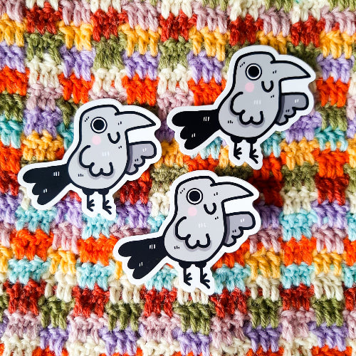 Kuvassa oleva vedenkestävä tarra on harmaa korppi, joka hymyilee iloisesti. Korpilla on suuret silmät ja pinkit, kirkkaat posket. Hauskan linnun peräsulat ovat oikein suuret ja komeat.
