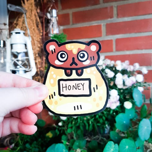 Kuvassa henkilön käsi pitelee magneettia. Magneetti on surullisen näköinen karhu, joka on jumissa hunajapurkissa. Karhu on ruskea ja sillä on hieman hunajaa päässä. Hunajapurkki on iso ja keltainen ja siinä lukee "honey" eli suomeksi "hunaja".