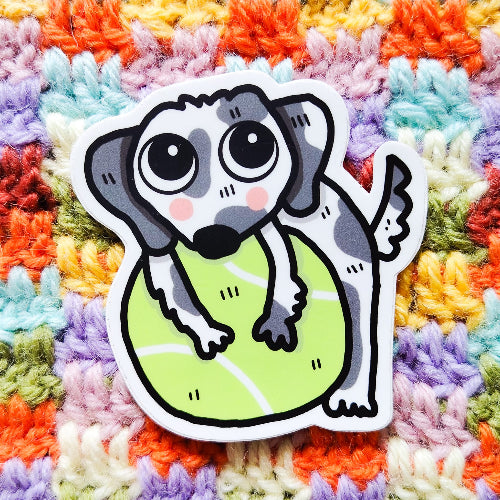 Kuvassa oleva tarra on dalmatiankoiran pentu halaamssa isoa, vihreää tennispalloa. Dalmatialainen tuijottaa katsojaa suurilla sympaattisilla silmillään.