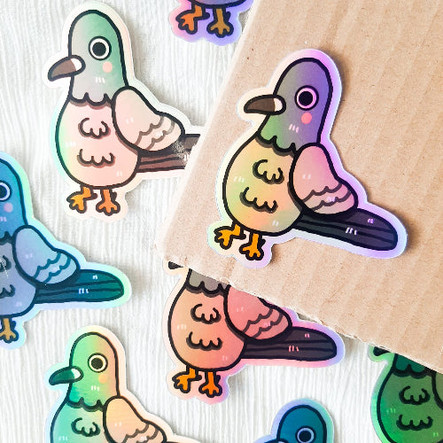 Kuvassa on pulutarra. Tarra on holograafinen, ja se loistaa sateenkaaren väreissä, kun siihen osuu aurinko. Pulu on harmaa, ja sillä on oikein komeat sulat, isot silmät ja vaaleanpunaiset posket.