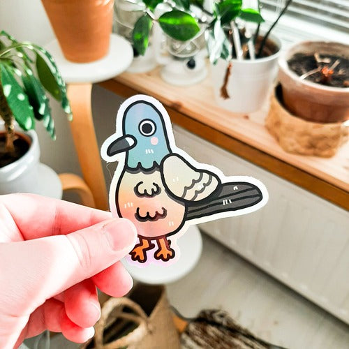 Kuvassa on pulutarra. Tarra on holograafinen, ja se loistaa sateenkaaren väreissä, kun siihen osuu aurinko. Pulu on harmaa, ja sillä on oikein komeat sulat, isot silmät ja vaaleanpunaiset posket.