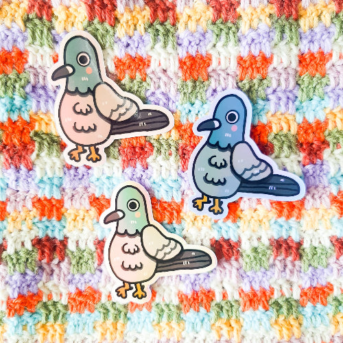 Kuvassa on pulutarra. Tarra on holograafinen, ja se loistaa sateenkaaren väreissä, kun siihen osuu aurinko. Pulu on harmaa, ja sillä on oikein komeat sulat, isot silmät ja vaaleanpunaiset posket.