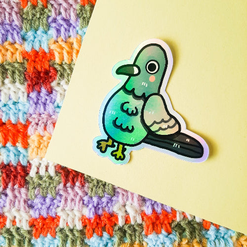 Kuvassa on pulutarra. Tarra on holograafinen, ja se loistaa sateenkaaren väreissä, kun siihen osuu aurinko. Pulu on harmaa, ja sillä on oikein komeat sulat, isot silmät ja vaaleanpunaiset posket.