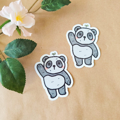 Kuvassa on hauska pandatarra. Panda on harmaavalkoinen, ja se nostaa toista tassuaan tervehdykseksi. Panda seisoo ja sillä on suloiset kiiluvat silmät. Sen päästä kasvaa pieni kasvi.