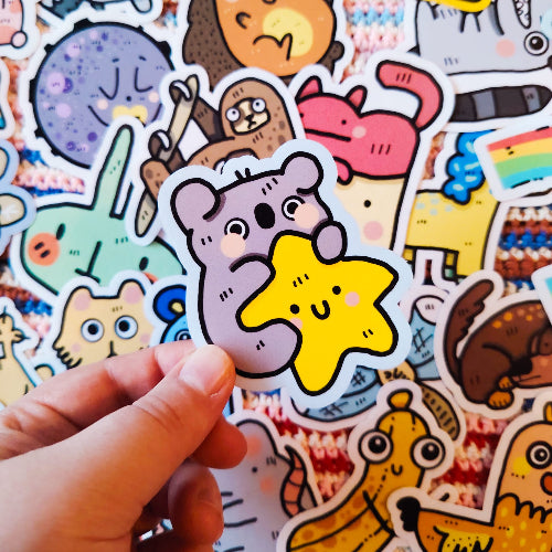 Kuvassa on käsi pitelemässä koalatarraa. Taustalla on paljon erivärisiä ja erilaisia eläintarroja