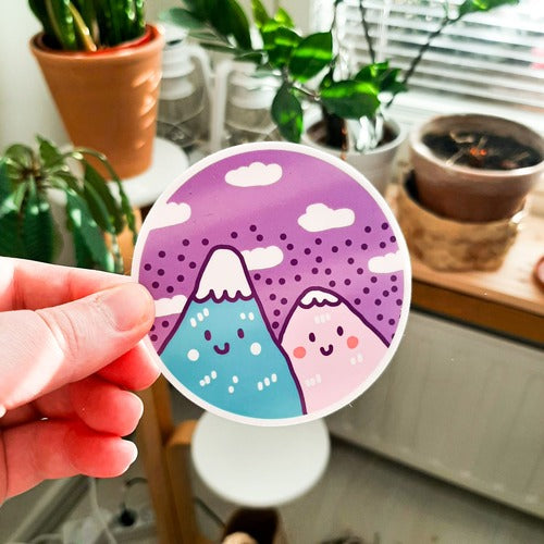 Kuvassa on henkilön käsi pitelemässä tarraa, jossa on kaksi vuorta. Tarra on pyöreä ja sen tausta on violetti. Taustalla on kodin huonekaluja ja kasveja.